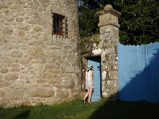 Un jeune femme entre par une porte bleue entourrée de puerres de tailles. La tour médiévale domine l'ensemble.