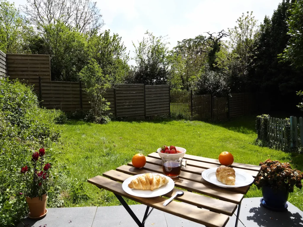 Une table de jardin est dressée avec un petit déjeuner. La vue s'ouvre sur une pelouse de 200 m².