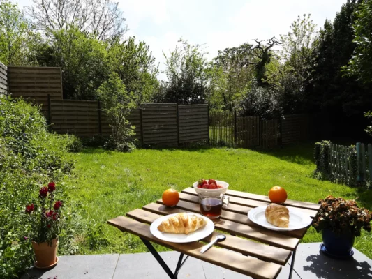 Une table de jardin est dressée avec un petit déjeuner. La vue s'ouvre sur une pelouse de 200 m².